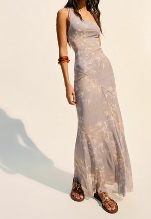 Robe longue et fluide en tissu léger et transparent avec un motif tie-dye lilas et beige. Elle possède de fines bretelles et une silhouette ajustée. Sandales marron.