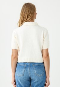 Weißer, kurzärmeliger Strickpullover mit Kragen und geripptem Saum, kombiniert mit hochgeschnittenen blauen Jeans. Weiche Textur und lässige Passform.