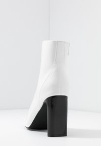 Weiße Knöchelstiefel mit schlankem Design, glänzendem schwarzen Blockabsatz, glatter Lederoberfläche und einem Reißverschluss auf der Rückseite.