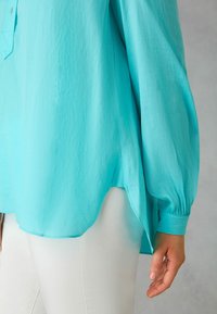 Blusa turchese realizzata in tessuto leggero, con maniche lunghe e gather, uno spacco laterale e una chiusura a bottoni sul colletto.