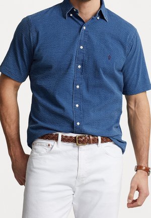 Mann, der ein kurzärmeliges, strukturiertes blaues Hemd mit Knöpfen trägt, weiße Hose, geflochtener brauner Gürtel und braune Lederuhr.