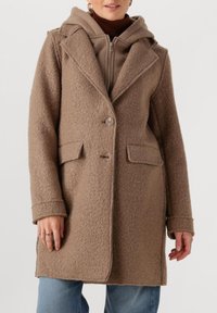 Vrouw in een bruine textuurwolleden jas over een bruine hoodie met ritssluiting, gecombineerd met blauwe jeans, staand tegen een effen achtergrond.