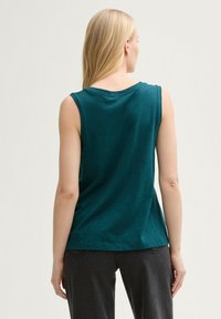 Top teal fără mâneci, fabricat dintr-un material moale, cu un guler rotund și o croială lejeră, asortat cu pantaloni gri închis.