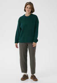 Grüner Strickpullover mit einem runden Ausschnitt, kombiniert mit hellbraunen karierten Hosen und braunen Schnürschuhen. Weicher Stoff, lockerer Schnitt.