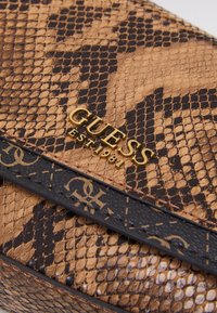 Ruskea ja musta käärmeennahkakuosiset Guess-lompakko, jossa on kultainen "GUESS EST 1981" -logo ja musta nahkareunus, jossa on monogrammitietoa.