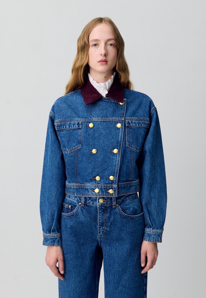 Giacca di jeans blu con design doppiopetto, bottoni dorati e colletto in velluto a coste bordeaux. Presenta tasche frontali e maniche dritte.