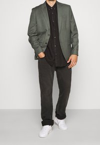 Blazer verde oliva, camicia nera con bottoni, pantaloni scuri, sneakers bianche. Caratteristiche includono due bottoni sul blazer e risvolti sui pantaloni.
