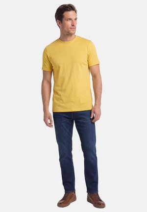 Mann steht, trägt ein gelbes kurzärmeliges T-Shirt, blaue Jeans und braune Sneakers, schaut nach links vor einem schlichten Hintergrund.