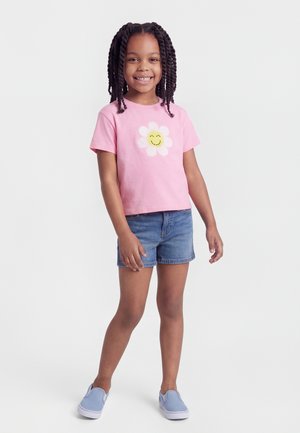 Smiling girl with braided hair wearing a pink t-shirt with a smiling flower and denim shorts, standing against a plain background.