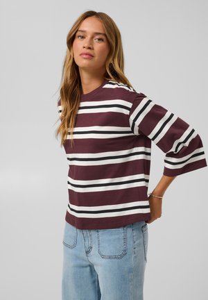 Mulher de cabelo comprido vestindo uma camisola às riscas em bordô, branco e preto e jeans azul claro, em pé com uma mão atrás das costas.