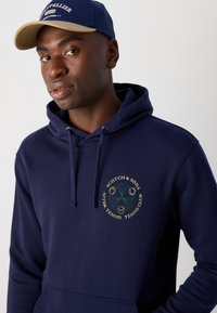 Sweat-shirt marine avec poche kangourou et capuche à cordon. Il présente un logo circulaire en jaune et vert, illustrant des éléments de tennis et des motifs de feuilles.