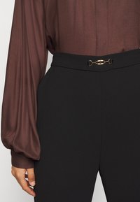 Blouse marron à manches longues rentrée dans un pantalon noir taille haute avec un petit détail de chaîne dorée à la taille, main reposant sur le côté.