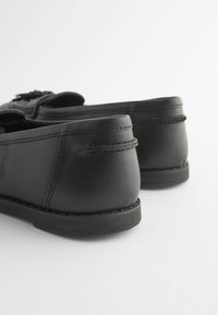 Mocasini din piele neagră cu o finisare netedă, vârf rotunjit și detalii de cusătură, având o talpă joasă din cauciuc și un ciucure decorativ în față.