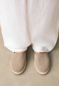 Beige mokkasiinit, joissa on juuttinauhasohva, yhdistettynä löysiin valkoisiin pellavahousuihin, nähtynä ylhäältä neutraalilla lattialla.