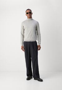 Pulover gri cu guler înalt, pantaloni negri cu dungi, și pantofi negri. Modelul poartă ochelari de soare verzi, stând pe un fundal alb.