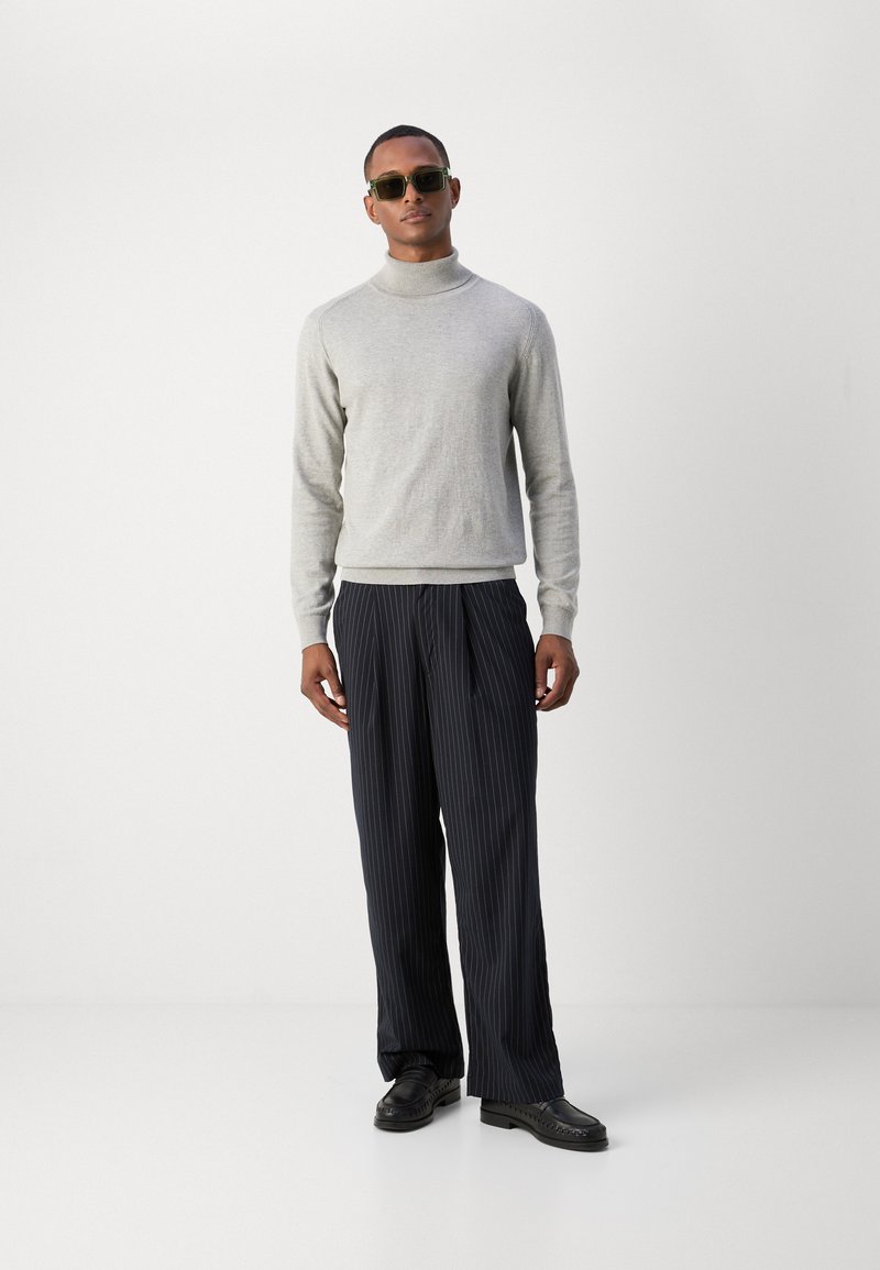 Pull en col roulé gris, pantalon à rayures sombres et chaussures noires. Le modèle porte des lunettes de soleil vertes et se tient sur un fond blanc.