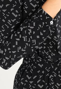Czarna bluzka z tkaniny z białymi wzorami w kropki i linie; posiada mankiet zapinany na guziki oraz marszczony, elastyczny pas dla dodatkowej tekstury i detalu wzorniczego.