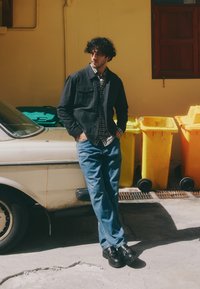 Črna kavbojka čez karirasto srajco, modri jeans in črne čevlje s debelim podplatom. Naslonjen ob avto blizu rumenih smetnjakov.