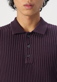 Fialová polokošile s žebrovanou strukturou a límečkem, opatřená légou se čtyřmi černými knoflíky. Texturovaná látka dodává hloubku.