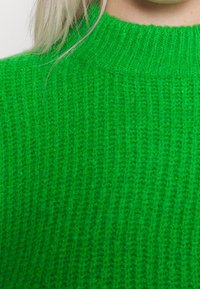 Zelený pletený svetr s texturovaným žebrovaným vzorem. Vysoký límec, měkký materiál, který zobrazuje pouze část oděvu.