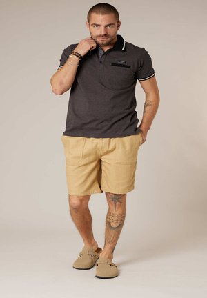 Homme portant un polo sombre, un short beige et des chaussures sans lacets, debout avec une main dans la poche et l'autre près du col.