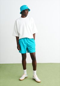 Camisa blanca de gran tamaño con mangas cortas, pantalones cortos turquesa brillante, gorra de cubo a juego, calcetines blancos y zapatillas de colores sobre un suelo verde.