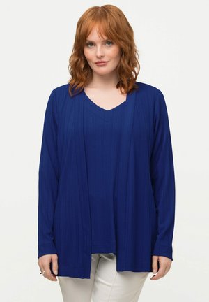 Langärmlige blaue Strickjacke mit strukturiertem, geripptem Design und V-Ausschnitt, über einem passenden Oberteil getragen, kombiniert mit hellfarbigen Hosen.