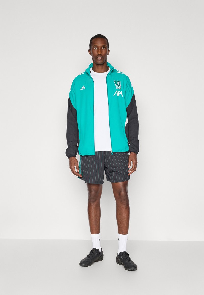 Veste zippée bleu-vert et noire avec logo, t-shirt blanc, shorts noirs à motifs, et chaussures de sport noires avec chaussettes blanches.