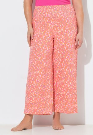 Vrouw draagt hoge taille, wijde broek met roze en oranje luipaardprint, staat blootsvoets tegen een witte achtergrond.
