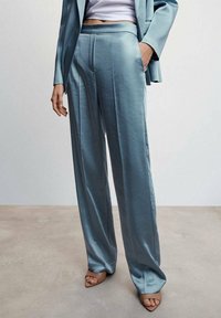 Hellblaue Satin-Hosen mit weitem Bein, sichtbaren Falten und glatter Textur. Kombiniert mit einem taillierten weißen Oberteil und neutralen Sandalen.