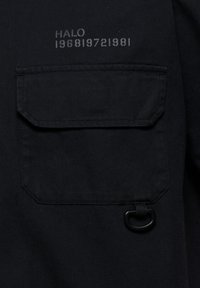 Schwarze Stofftasche mit Klappe, darüber bedruckter Text "HALO 1968|972|981" und unterhalb der Tasche ein kleiner schwarzer D-Ring befestigt.