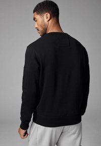 Sort bomuldssweatshirt med rund halsudskæring og ribbede manchetter, som har en firkantet patchdetalje på den øvre ryg og et minimalistisk design.