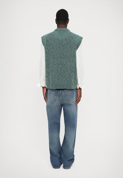 Groen gemêleerd mouwloos vest gemaakt van textuurstof, gedragen over een witte blouse met lange mouwen. Blauwe wijde spijkerbroek maakt de look compleet.