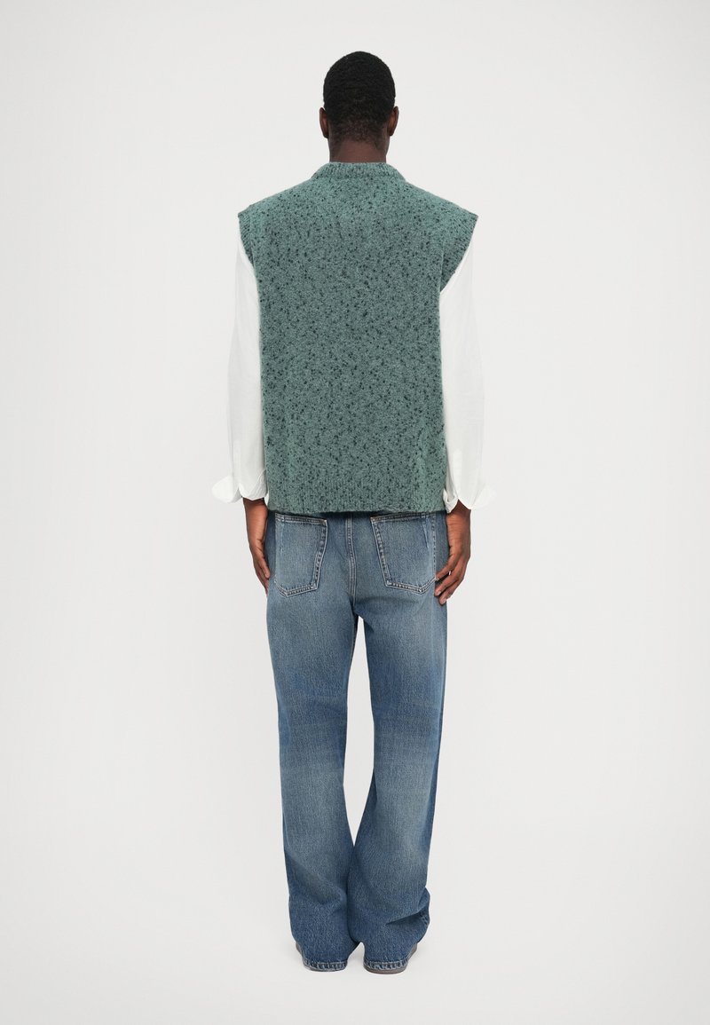 Gilet senza maniche verde maculato realizzato in tessuto testurizzato, indossato sopra una camicia bianca a maniche lunghe. Pantaloni in denim a gamba larga di colore blu completano il look.