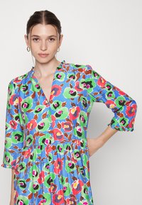 Vestido com estampa floral apresentando um fundo azul, tons de verde, rosa e castanho, com decote em V e mangas com fendas. Tecido leve.