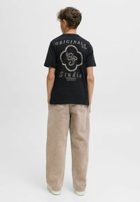 Črna bombažna majica s potiskom na hrbtni strani, ukrivljenim v zlatem tisku. Ohlapno prileganje, kombinirano s bež širokimi hlačami in belimi podplati.