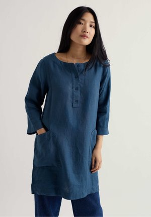 Mujer con cabello largo y negro, vistiendo una túnica azul de manga larga con botones y bolsillos frontales, de pie frente a un fondo claro y simple.