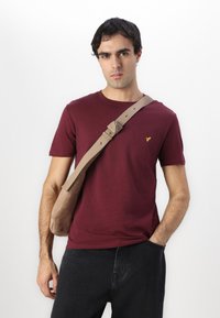 Burgundy short-sleeve t-shirt with a chest pocket featuring a yellow logo detail. The model also wears a beige shoulder bag with a textured strap.