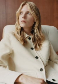 Giacca color crema in misto lana con bottoni neri, con collo e taschino sul petto, indossata con capelli lunghi e mossi su uno sfondo neutro.