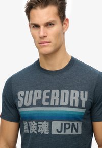 Camiseta de manga corta azul marino hecha de algodón, que presenta un texto en negrita "SUPERDRY" en gris con un diseño de rayas azules y caracteres japoneses.
