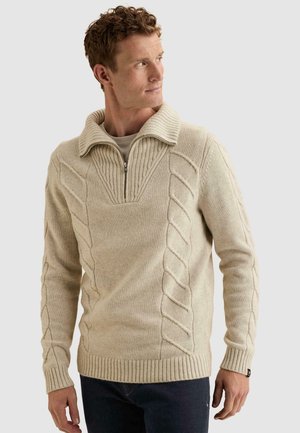 Hombre con jersey beige de punto trenzado con cremallera y cuello, mirando hacia un lado, sobre un fondo liso gris claro.