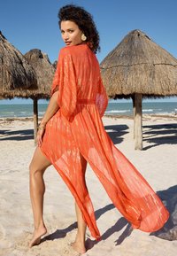 Cubierta ligera en color naranja con un diseño etéreo y fluido, y detalles de franjas doradas. Presenta mangas sueltas y una cintura atada. Entorno de playa.