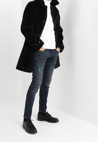 Svart, lång kappa i fluffigt tyg med krage och knappar, kombinerad med åtsittande jeans och svarta snörskor. Vit polotröja under.