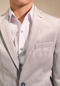 Hellgrauer Samtblazer mit strukturierter Gestaltung, einer Brusttasche und blauen Knöpfen, kombiniert mit einem knusprig weißen Hemd.