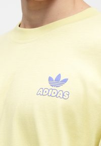 Ανοιχτό κίτρινο βαμβακερό μπλουζάκι που διαθέτει το μπλε λογότυπο της Adidas με τρεις ανοιξιάτικες λεπτομέρειες. Στρογγυλή λαιμόκοψη και απαλό υλικό.