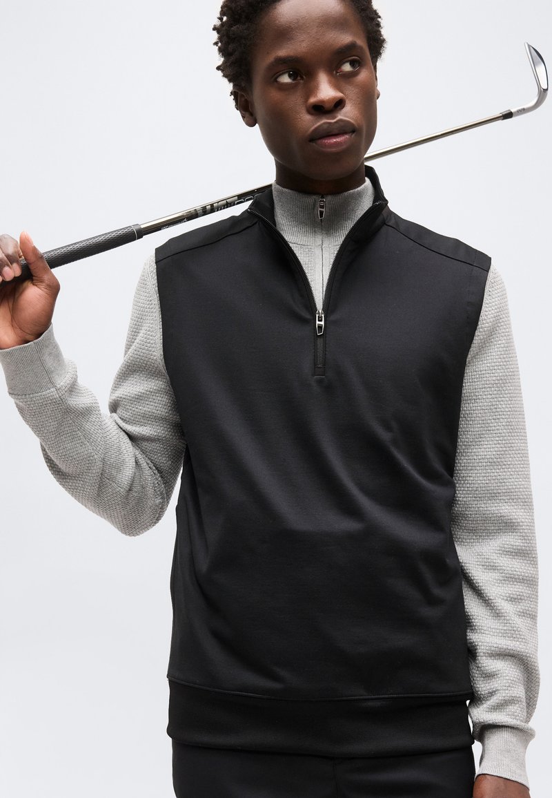 Jeune homme portant un gilet noir sans manches à fermeture éclair par-dessus une chemise grise à manches longues, tenant un club de golf sur son épaule.