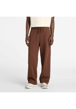 Person trägt lockere braune Hose mit Kordelzug und weiße Turnschuhe und steht auf einem schlichten hellen Hintergrund.