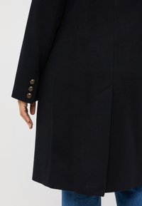 Fekete gyapjú kabát hosszú szsilhuettel, három díszgombbal az ujjon és oldalsó hasítékkal a könnyebb mozgás érdekében.