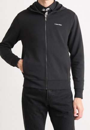 Mann trägt schwarzen Calvin Klein Zip-Hoodie über einem gemusterten Hemd, eine Hand in der Tasche, vor neutralem Hintergrund.