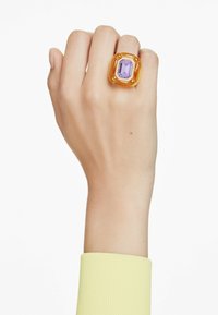 Grande anello arancione con un gemma viola a taglio smeraldo circondata da piccole pietre chiare; indossato su una mano con una manica gialla a righe.